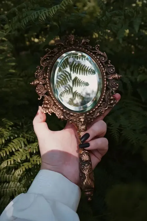 Una mano regge uno specchio antico che riflette le felci del sottobosco